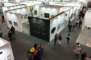 People browsing artwork at a contemporary art gallery with various paintings and installations displayed on white walls.