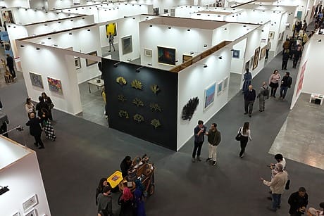 People browsing artwork at a contemporary art gallery with various paintings and installations displayed on white walls.