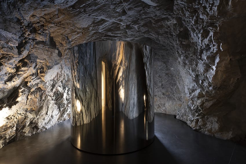 Sleek mirrored cylinder in a rocky cave with textured walls and dim lighting, creating a dramatic and modern aesthetic.