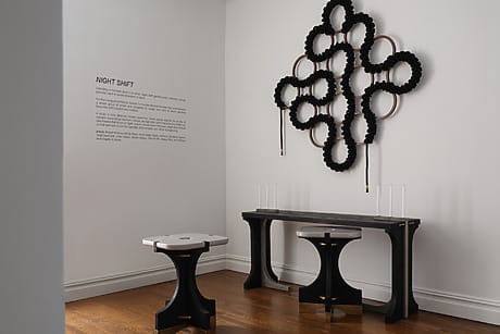 modern art installation with black sculptures on a white wall, wooden floor, three dark furniture pieces, and a text panel
