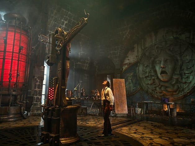 A person stands in a gothic, steampunk room with a large red cylinder and Medusa face on the wall.