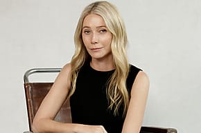 Person with long blonde hair sitting on a chair, wearing a black sleeveless top, against a plain background.