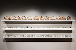 Three white shelves displaying wooden twist sculptures on top and various small, colorful objects on the lower two shelves.