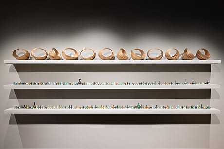 Three white shelves displaying wooden twist sculptures on top and various small, colorful objects on the lower two shelves.