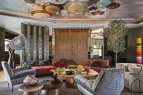 Eclectic living room with colorful ceiling art, modern furniture, and decorative accents around a central coffee table.