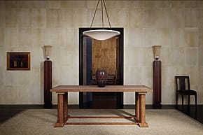Minimalist dining room with a wooden table, decorative vase, two tall lamps, a painting, and a single chair against beige walls.