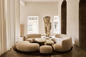 Minimalist room with curved beige sofas, round tables, and a headless statue by large windows.