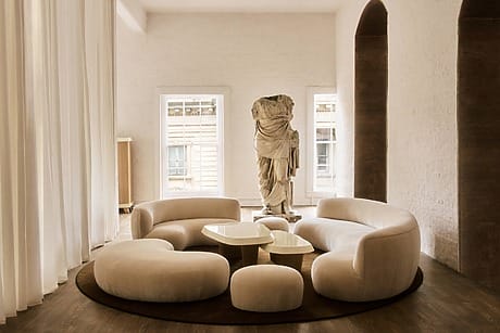 Minimalist room with curved beige sofas, round tables, and a headless statue by large windows.