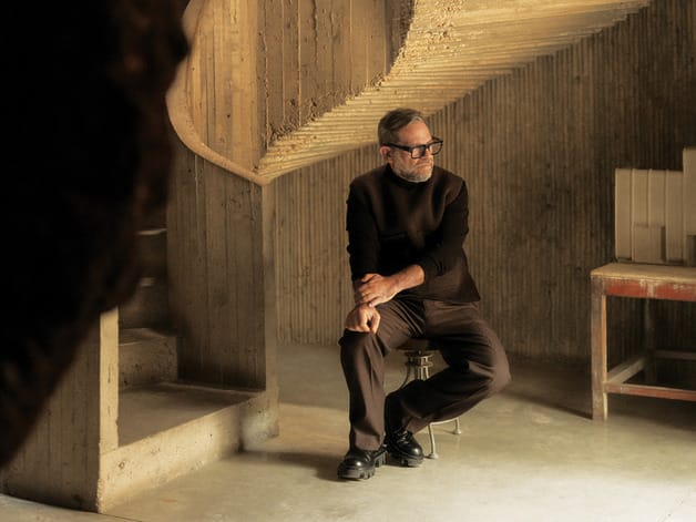 Man sitting on a stool in a dimly lit, textured concrete interior, wearing a dark outfit and glasses.