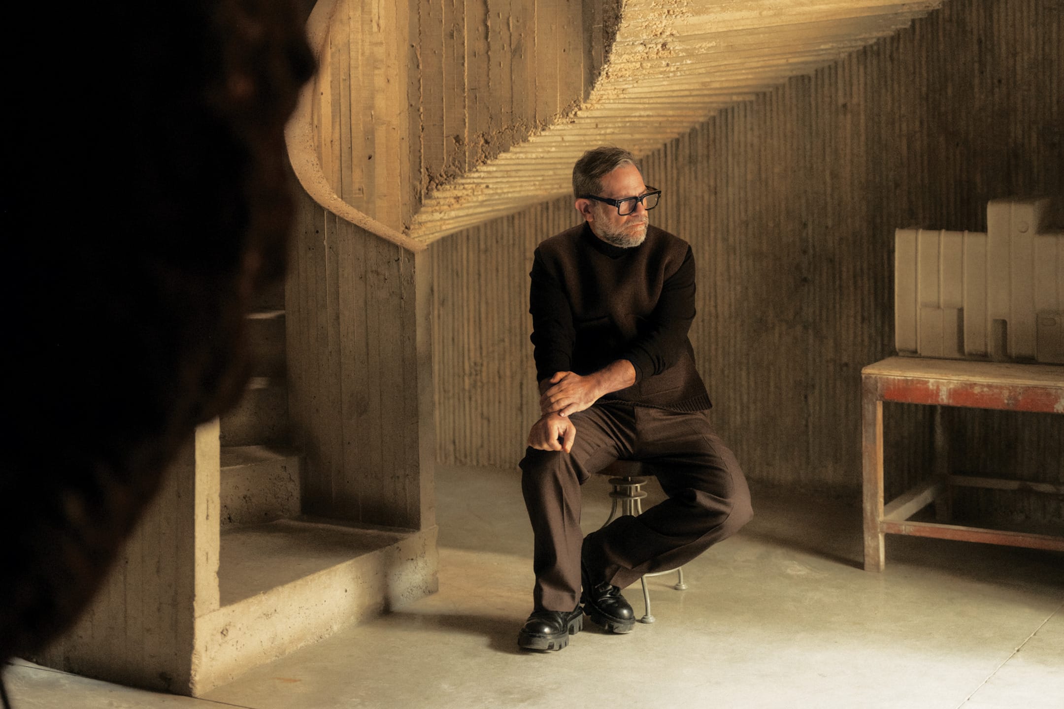 Man sitting on a stool in a dimly lit, textured concrete interior, wearing a dark outfit and glasses.