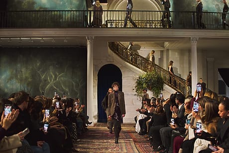 Model walking down fashion show runway with audience capturing photos on phones, stylish venue with elegant staircase background