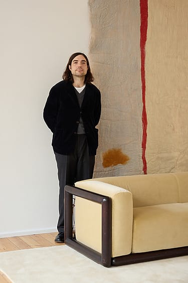 Person standing next to a modern beige sofa with a textured, abstract wall tapestry in the background.