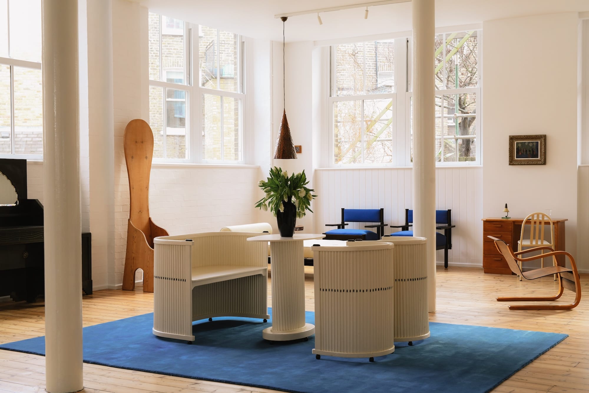 Modern office space with white rounded desks, blue rug, wooden flooring, and large windows allowing natural light in.