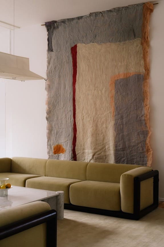 Abstract wall hanging with earthy tones behind two green sofas and a marble coffee table in a modern living room.