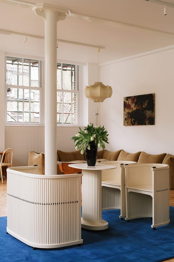Modern living room with blue rug, cream furniture, column, and vase of white flowers. Large window and couch in the background.