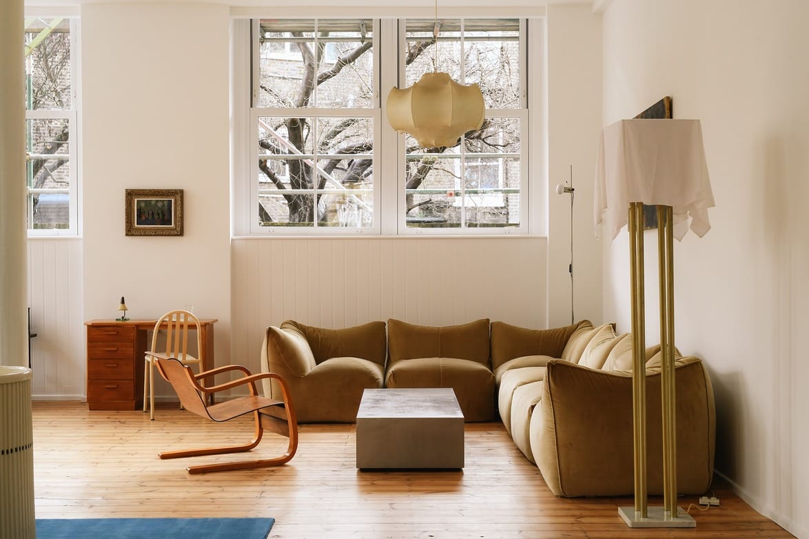 Minimalist living room with modern furniture, including a tan sofa, unique chairs, wooden desk, and tall windows.