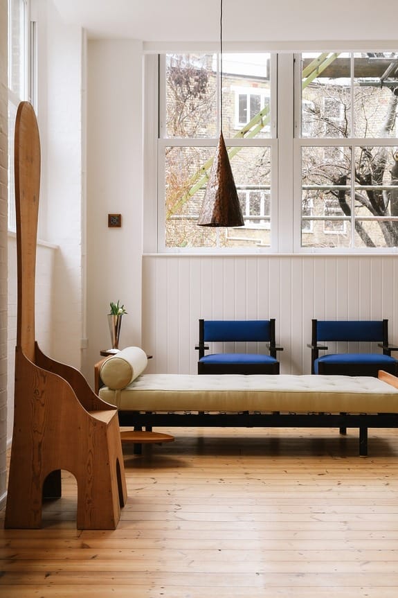 Modern living room with wooden floors, minimalist furniture, large windows, and unique wooden chair near a wall.