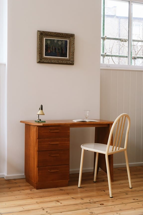 Wooden desk and chair in a minimalist room with a small lamp and a picture frame on the wall above.