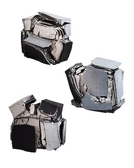 Three abstract sculptures made of layered materials in gray and black, resembling geometric shapes.