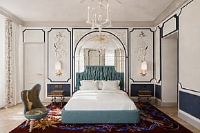 Luxurious bedroom with blue upholstered bed, ornate wall art, chandelier, and decorative chair on patterned rug.