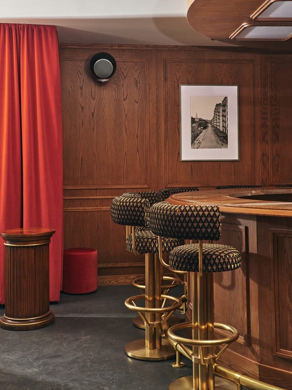 Stylish bar interior with wooden paneling, gold barstools, a red curtain, and a framed black-and-white photo on the wall.
