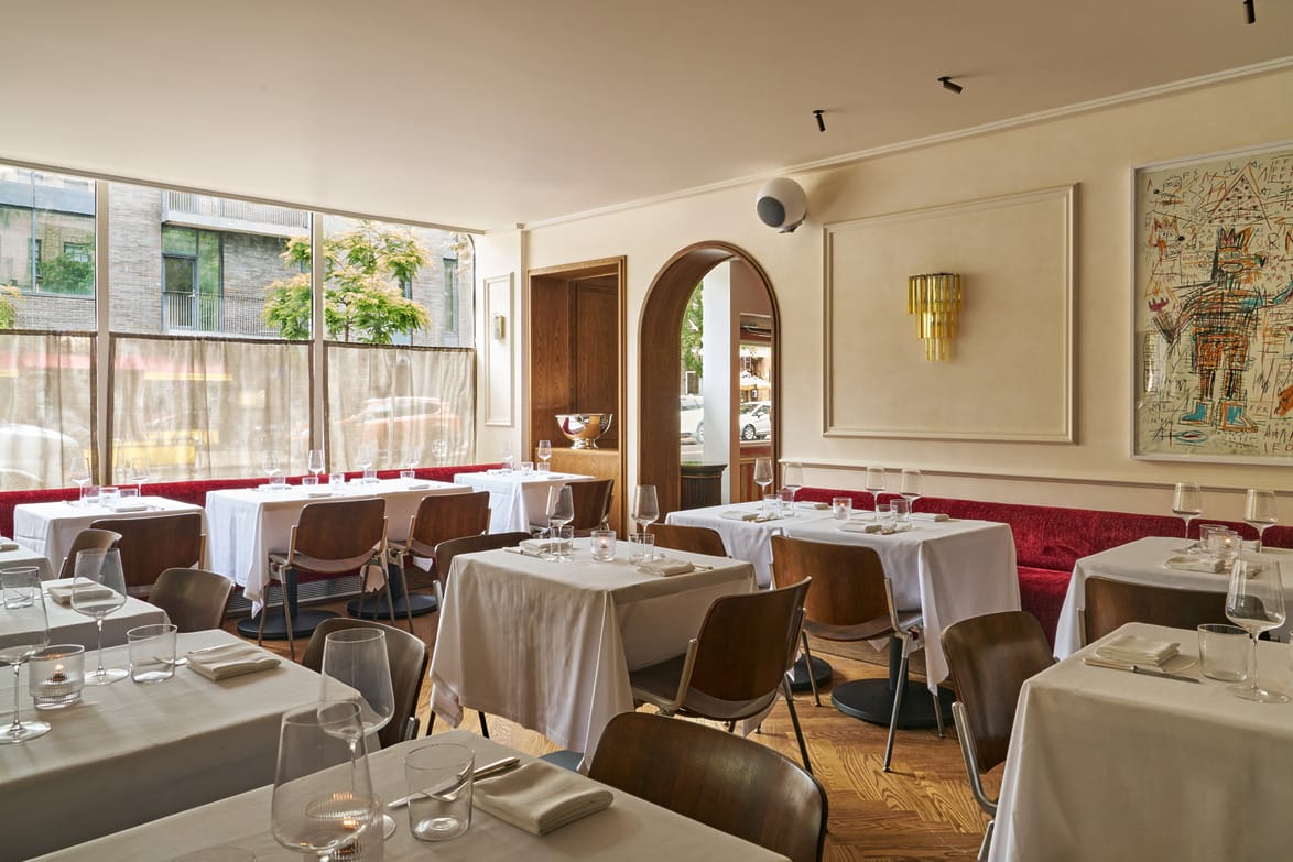 Cozy restaurant interior with white tablecloths, wooden chairs, large windows, and framed art on the walls.