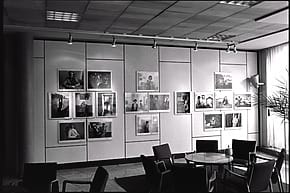 Black and white photo of a gallery wall displaying multiple framed photographs, with chairs and tables in the foreground.