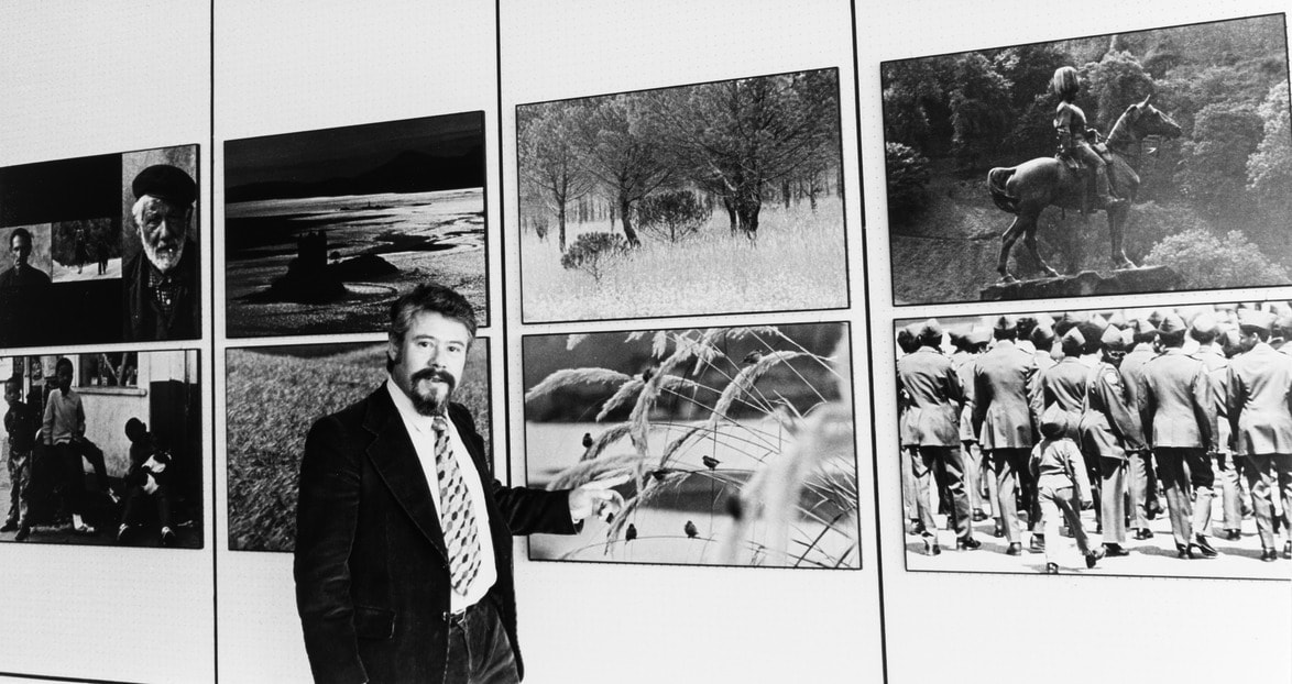 Man stands beside a display of black and white photographs, pointing towards the images, wearing a suit and tie.