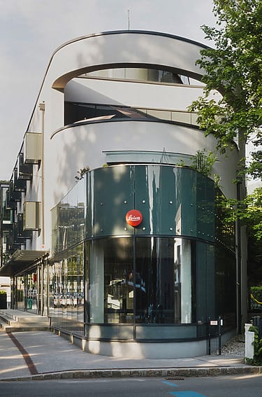Modern three-story building with curved facade, glass windows, and Leica logo, surrounded by trees and a sidewalk.