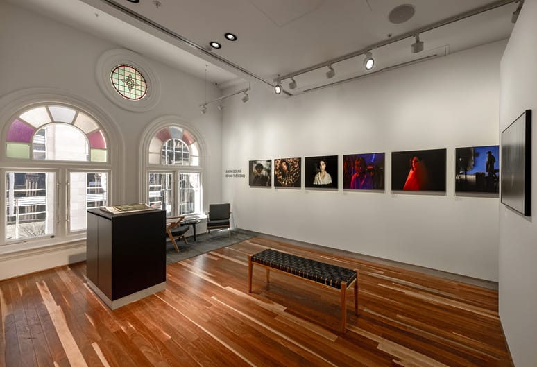 Art gallery room with wooden floors, a bench, photographs on walls, and arched windows with stained glass.
