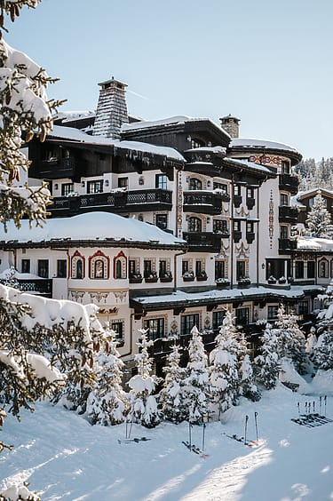 Snow-covered alpine hotel surrounded by pine trees and ski equipment under a clear blue sky