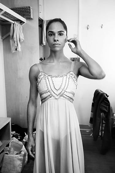 Ballerina in a dress adjusting hair in a dressing room, with clothes and a chair in the background.