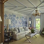 Elegant living room with patterned blue wallpaper, white sofa, decorative items, chandelier, and natural light from large windows.