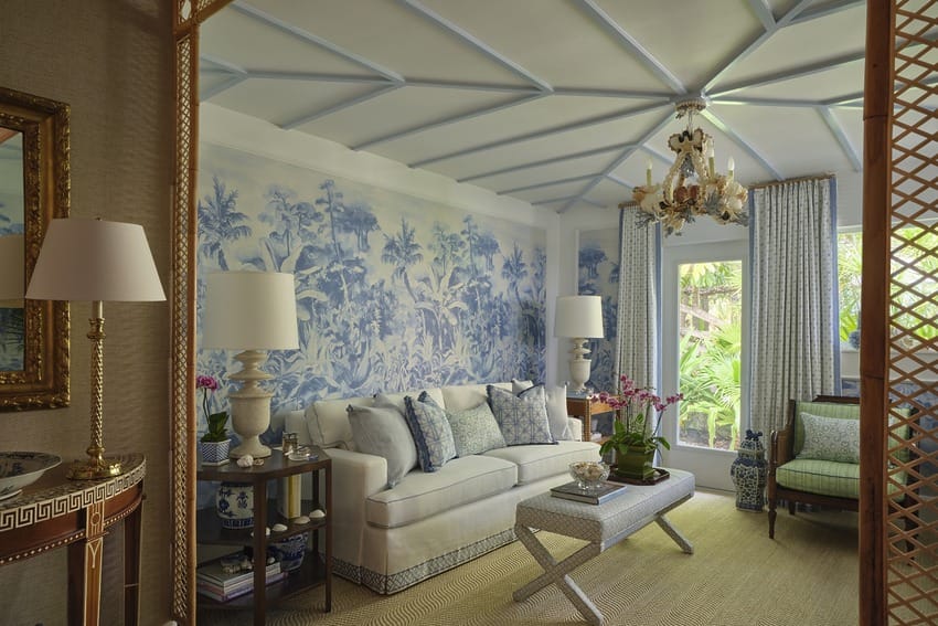 Elegant living room with patterned blue wallpaper, white sofa, decorative items, chandelier, and natural light from large windows.