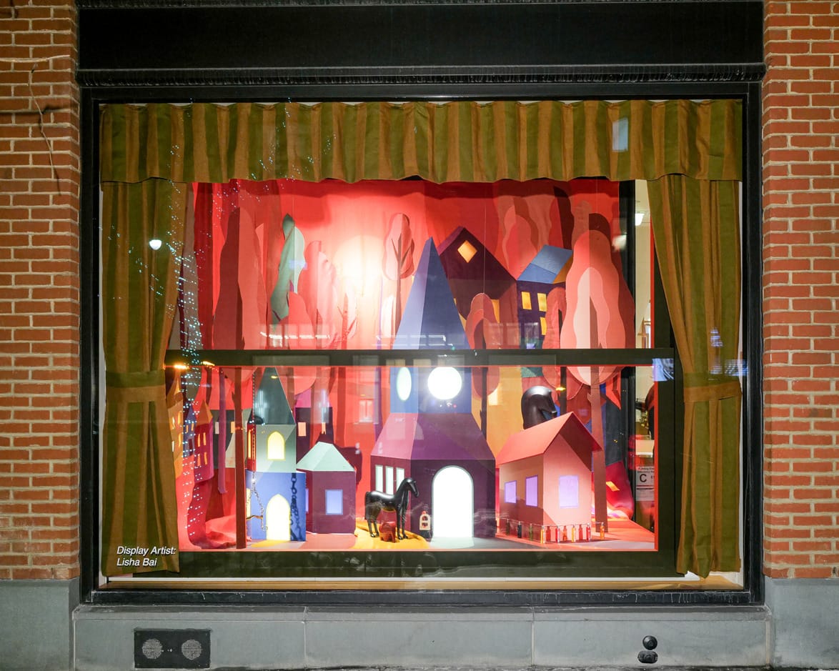 Storefront window display featuring a colorful paper village with illuminated houses and trees, decorated in vibrant hues.
