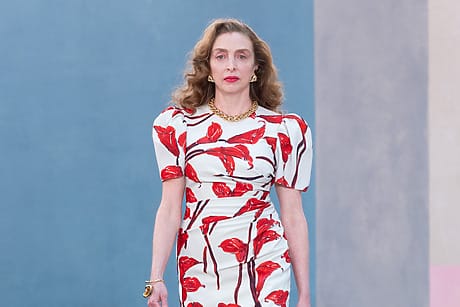 Model walking in a fashion show wearing a white dress with red floral prints and red high heels.