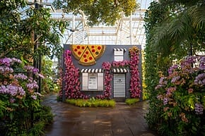 Botanical garden with a small brick building adorned with floral displays and a large pizza slice decor.