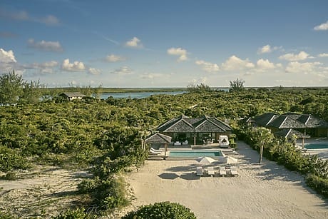 Aerial view of a tropical beach resort with villas, pools, sandy shore, and lush greenery under a clear blue sky.