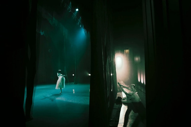 Ballet dancer performing gracefully on stage with dim lighting, while another dancer waits backstage.