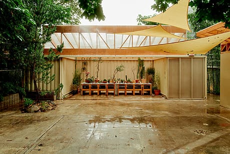 Outdoor patio with wooden tables, chairs, and potted plants under shade sails, surrounded by trees and greenery.