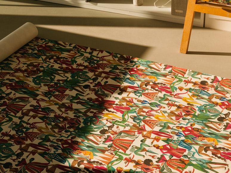 Patterned fabric with colorful abstract figures rolled out on a floor, sunlight casting shadows across the surface.