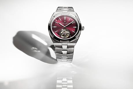 Luxury wristwatch with a metallic band and a red dial placed on a reflective surface, casting a shadow on a white background.