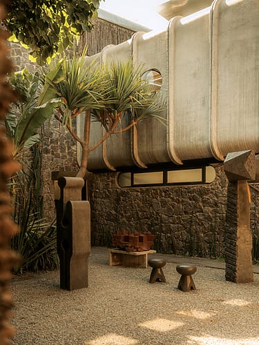 Modern outdoor space with sculptural elements, stone walls, and tropical plants under a covered walkway.