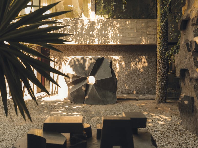 Outdoor sculpture garden with large abstract stone centerpiece surrounded by plants and warm sunlight filtering through leaves