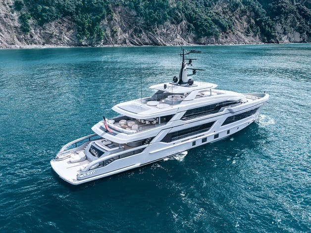 Luxurious white yacht sailing in clear blue waters near a rocky coastline with lush greenery above.