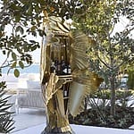 Artistic bird-shaped bar cabinet outdoors with wine and glasses, surrounded by greenery and overlooking a body of water.