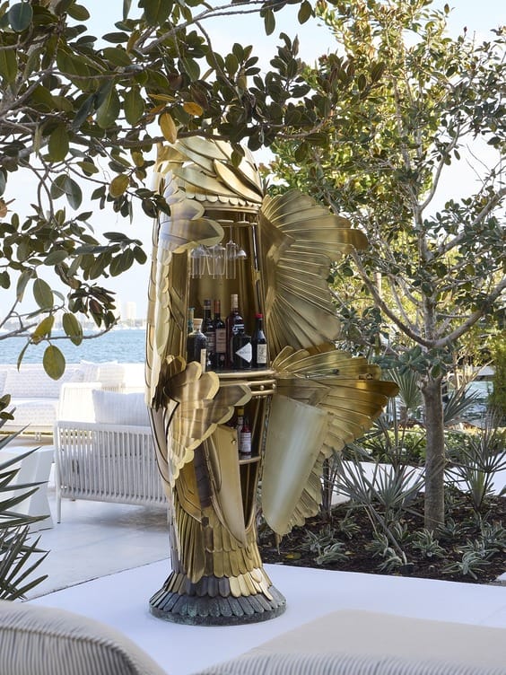 Artistic bird-shaped bar cabinet outdoors with wine and glasses, surrounded by greenery and overlooking a body of water.