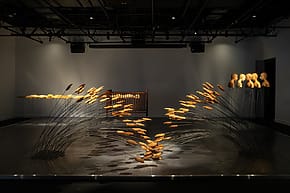 Modern art installation with abstract sculptures and curved metal rods, displayed in a dimly lit gallery space.