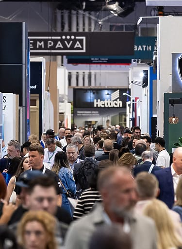 Crowded exhibition hall with people walking and various brand signs visible overhead.