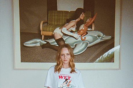 Woman in white shirt stands in front of large artwork featuring a woman with inflatable dolphin and chair.